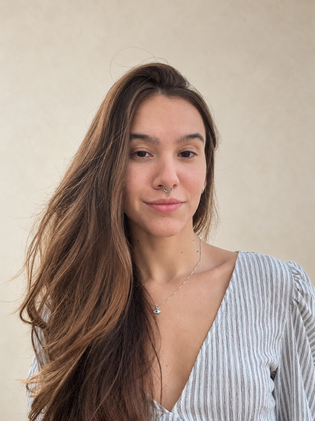 Letícia Paola com cabelos longos castanhos, blusa listrada e colar, em uma fotografia com fundo neutro claro.